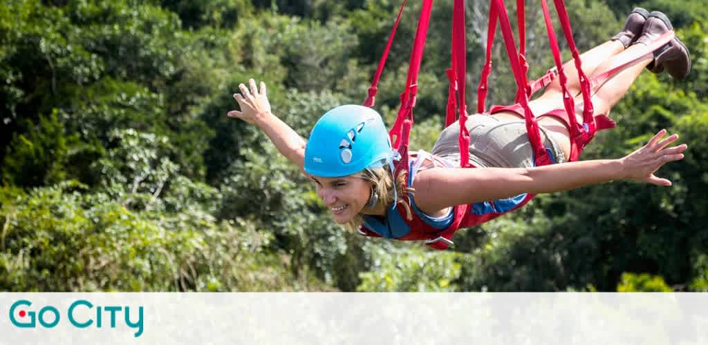 A person with a blue helmet is enjoying a thrilling zipline adventure through lush green trees. They are wearing a harness and are spread out in a flying position with a big smile. The background is filled with dense foliage. Go City logo is at the bottom left.