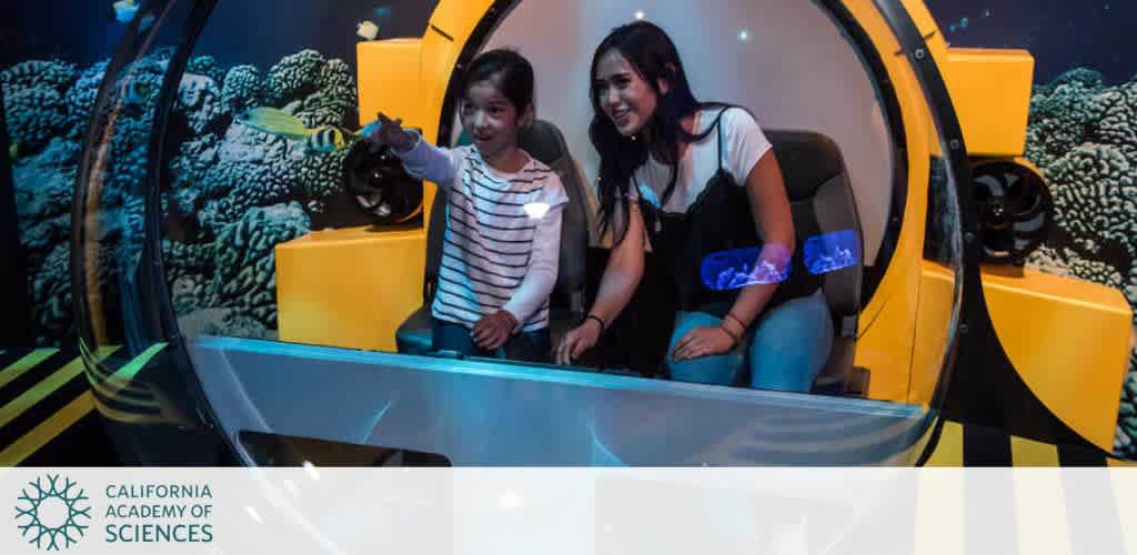 Two people inside a submarine exhibit capsule pointing at an underwater display.