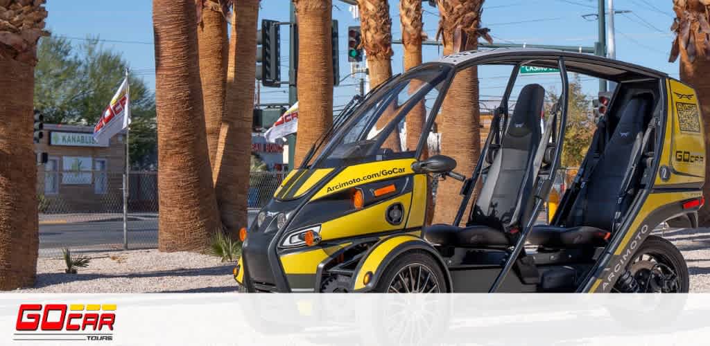 This image features a bright yellow and black three-wheeled GoCar parked on an urban street, awaiting adventurous riders. The compact, two-seater vehicle is designed with an open side structure, offering an unobstructed view of the surroundings to its passengers. In the background, one can see a row of tall palm trees lining the street, blending with the clear blue sky. A fence with flags is partially visible behind the trees, displaying a mix of red and white colors that add a vibrant touch to the scene. The GoCar’s branding is prominently displayed on both the vehicle and a logo watermark in the lower left corner, highlighting its presence as a tour service. At GreatWorkPerks.com, we're dedicated to ensuring you have an exhilarating experience with the added benefit of securing your tickets at some of the lowest prices available, guaranteeing significant savings on your next adventure.