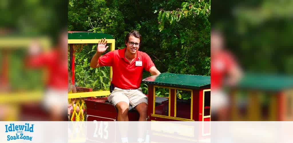 A person wearing a red shirt and khaki shorts is happily waving while sitting on a small train. The train is colorful with a green and red engine. Lush green trees are visible in the background, creating a lively and fun atmosphere. The logo of Idlewild and SoakZone is at the bottom left corner of the image.
