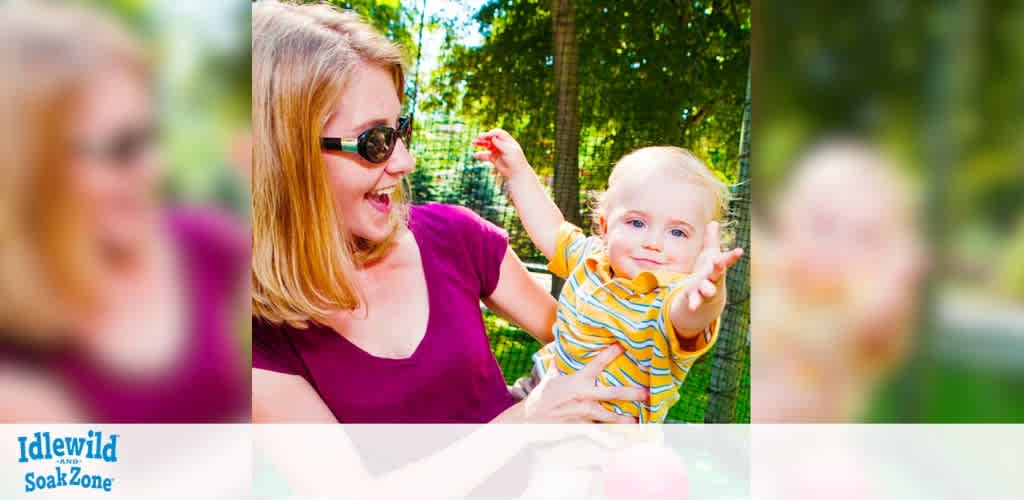 A smiling woman wearing sunglasses joyfully lifts a baby in a playful outdoor setting. The baby, dressed in a yellow striped shirt, extends their arms happily. Sunlight filters through the trees, creating a warm and cheerful atmosphere. The Idlewild and SoakZone logo is visible in the bottom left corner.