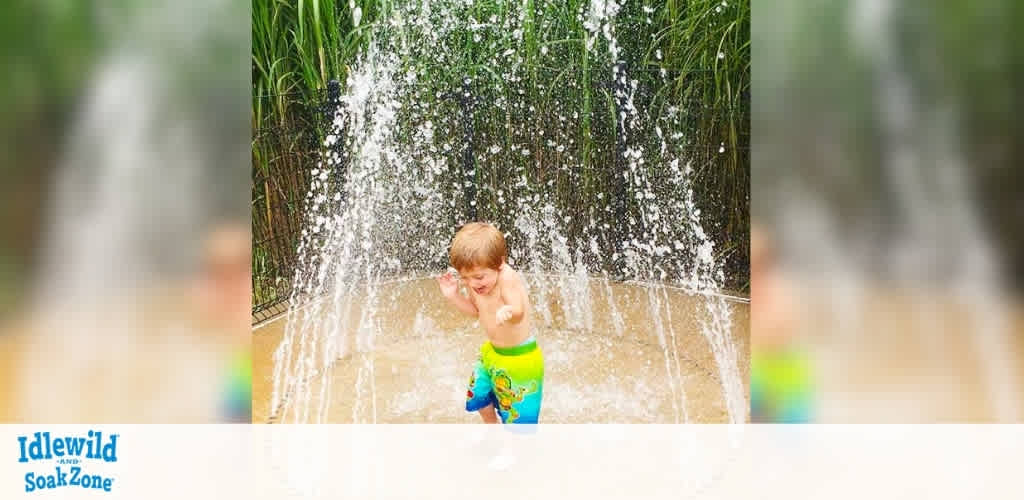 A young child enjoys a splash pad, standing under a curtain of water streams. The kid is wearing colorful swim trunks and appears to be delighted by the water. Tall green plants surround the background, adding to the playful outdoor setting. The atmosphere is lively and fun, ideal for a refreshing summer activity. Idlewild SoakZone logo is visible at the bottom left of the image.