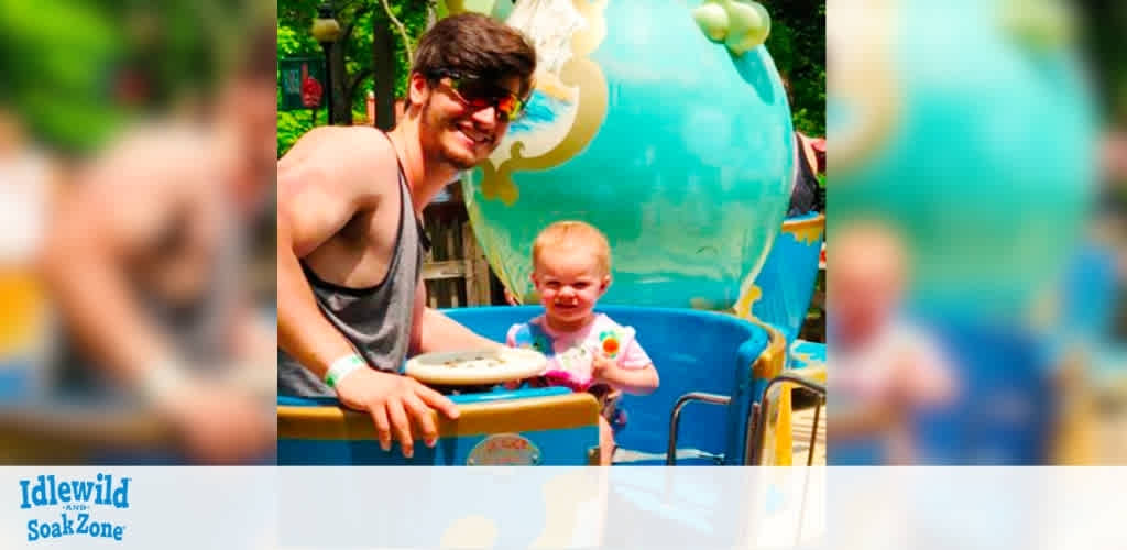 A man and a young child enjoy a sunny day at Idlewild and Soak Zone. They are seated in a colorful ride shaped like a teacup, surrounded by lush greenery. The man smiles, wearing sunglasses and a tank top, while the child looks happily at the camera. The vibrant colors and cheerful atmosphere capture the fun and excitement of a day at the amusement park.