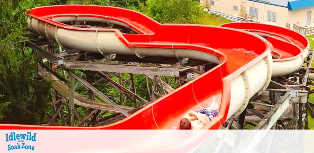 A person slides down a twisting red water slide at Idlewild and SoakZone. The slide winds through lush green trees, providing an exhilarating outdoor water adventure. The sturdy wooden support structures of the slide are visible, adding to the rustic amusement park feel. The bright colors and surrounding greenery create a vibrant, fun atmosphere for visitors seeking excitement and relaxation.
