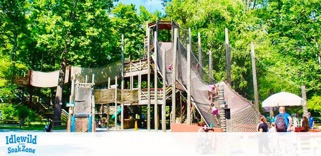 The image shows a vibrant outdoor playground surrounded by lush green trees, part of Idlewild and SoakZone. It features climbing structures and net bridges with families and children playing. The sunny day highlights the fun and adventurous atmosphere of the park, creating an inviting environment for all ages to explore and enjoy.