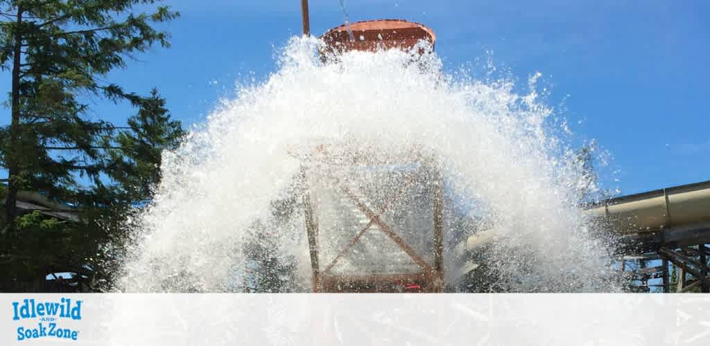 A massive splash of water cascades down from a tall wooden structure at a water park. The bright blue sky and adjacent water slides frame the scene. Lush green trees are visible on the left, adding natural beauty to the lively outdoor setting, which is Idlewild and SoakZone.