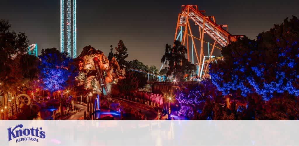 Evening view of Knott's Berry Farm highlighting the illuminated roller coaster tracks and landscape lighting. Trees glow with blue lights while orange-lit coaster structures rise against the night sky.