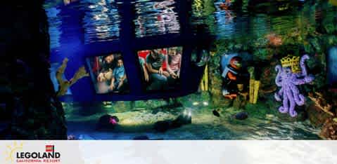 Image depicts an underwater-themed LEGOLAND California Resort attraction. Visitors are seen inside a transparent submarine-like vehicle observing colorful marine life, including a purple octopus figure and various fishes, made from LEGO bricks in a vibrant underwater setting. The LEGOLAND logo is visible in the bottom right corner.