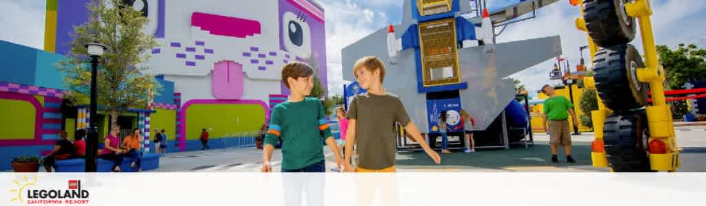 This image features a vibrant scene at LEGOLAND California Resort with visitors walking and conversing. Brightly colored buildings with blocky designs reminiscent of LEGO bricks line the background. A young boy in the foreground appears to be in mid-conversation with another, against a backdrop of an oversized LEGO vehicle model and sunny blue skies.