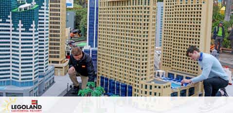 Two individuals are engaged in the maintenance of a LEGO display featuring intricate models of skyscrapers, focusing on the arrangement at LEGOLAND. Greenery is being placed by one of the workers, contributing to the vibrant miniature cityscape set against a LEGOLAND backdrop.