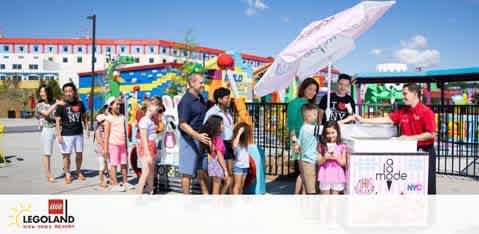 Visitors line up under a sunny sky outside Legoland. A group of children and adults await their turn at a booth with a staff member. Brightly colored Legoland structures are in the background, and promotional banners for Legoland and Madame Tussauds New York are visible.