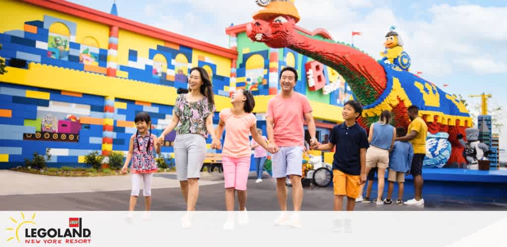 An image featuring a family smiling and holding hands at LEGOLAND New York Resort. They walk by vibrant Lego brick structures, including a large red dragon, with visitors around. The LEGOLAND logo is visible below.