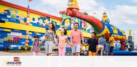 Image shows a happy group of visitors, including adults and children, walking in front of a colorful LEGOLAND entrance adorned with large LEGO sculptures, including an oversized red LEGO dragon overhead. Clear skies with few clouds set a cheerful atmosphere for a fun day out.