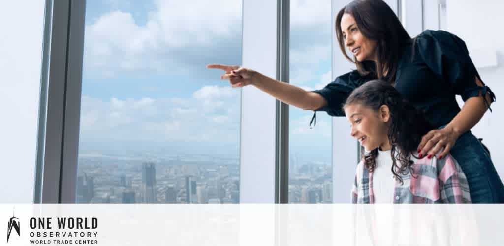 Image shows a woman and a young girl at the One World Observatory. They are indoors, looking out large windows at a cityscape. The woman, standing, points out something in the distance, while the girl, in front of her, looks on with interest.