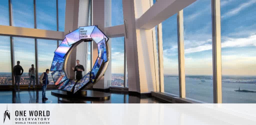 Inside the One World Observatory, visitors stand by large windows offering panoramic views of the cityscape and waterfront. A prominent, illuminated display in the shape of the number '0', with the text 'One World Trade Center' inside, is the centerpiece. The sky is clear, hinting at a high vantage point above the city.