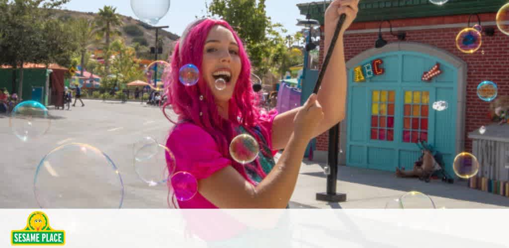 Person with pink hair playing with bubbles outside a colorful building.