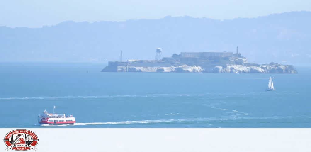 View of Alcatraz Island with a red and white ferry and a white sailboat in the foreground on San Francisco Bay.