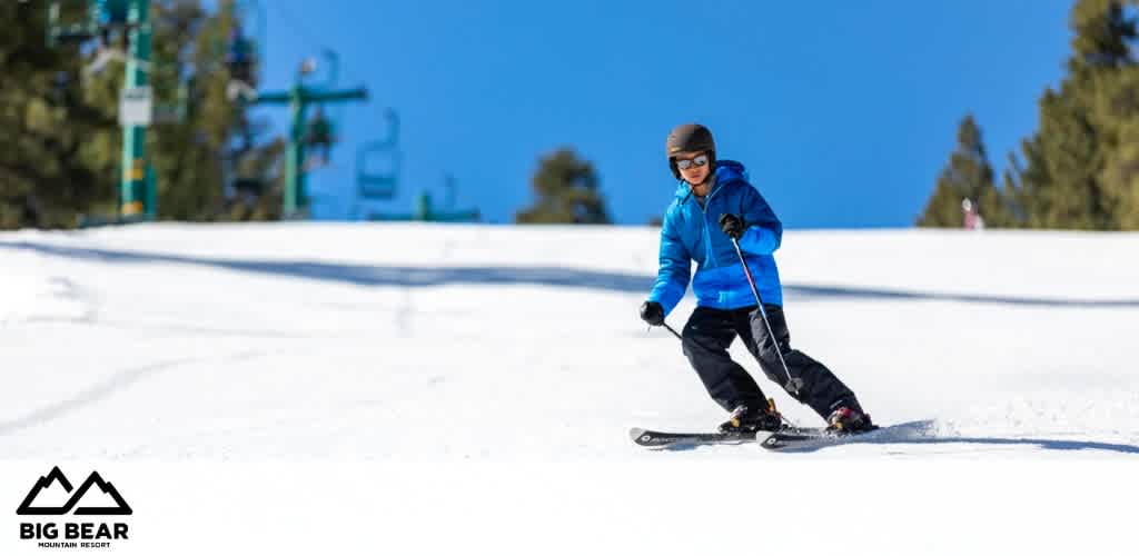 This image showcases an individual skiing down a gentle slope at Big Bear Mountain Resort under a clear blue sky. The skier, positioned to the right of the frame, is dressed in a bright blue jacket, dark pants, and is wearing a helmet and goggles for safety. The snow-covered slope appears well-groomed, and in the background, ski lifts and tall evergreen trees are visible, indicating an alpine environment. The Big Bear Mountain Resort logo is prominently displayed on the bottom left corner of the image.

At GreatWorkPerks.com, adventure awaits at the lowest prices – be sure to grab your discount tickets for thrilling experiences just like this one!