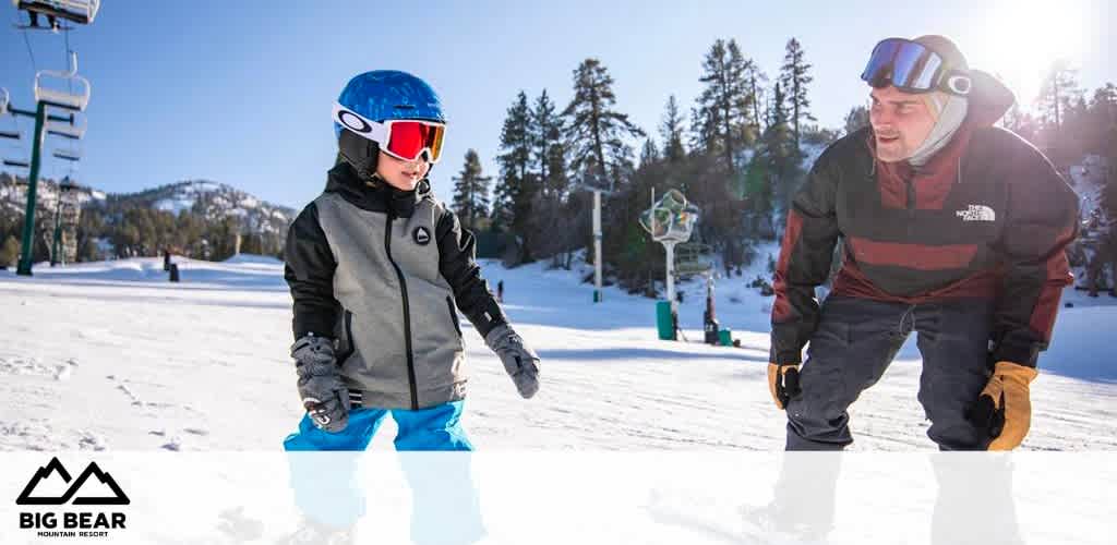 This image captures a joyful moment at the Big Bear Mountain Resort, with a clear blue sky above and snow-covered slopes in the background. On the left, a child wearing a helmet, goggles, and winter gear, stands confidently on the snow, facing the camera. The child is in the foreground and appears to be prepared for skiing or snowboarding, sporting a grey jacket with black and blue accents as well as blue snow pants. To the right, an adult male, who seems to be providing instruction or encouragement, leans slightly forward with a focus on the child. He is wearing a striped jacket, gloves, and a helmet with goggles pushed up onto the forehead, indicative of a casual teaching moment or shared activity. Both are situated at the base of a ski lift, with alpine trees and a clear view of the mountain in the backdrop, setting the scene for winter sports enthusiasts. This picture conveys the essence of winter fun and learning in a family-friendly environment.

At GreatWorkPerks.com, we believe in maximizing your snowy escapades with the lowest prices on tickets, ensuring substantial savings as you carve your path down the picturesque slopes of Big Bear Mountain Resort.