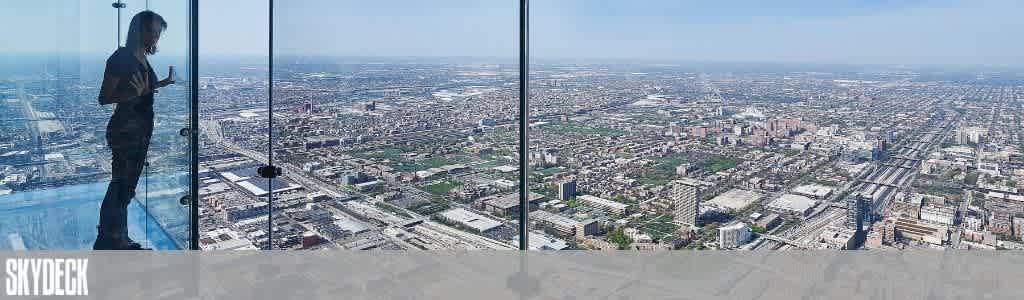 A person stands on a glass ledge at Skydeck, overlooking a vast urban landscape. The view showcases a sprawling city with numerous buildings and streets stretching into the horizon under a clear blue sky.