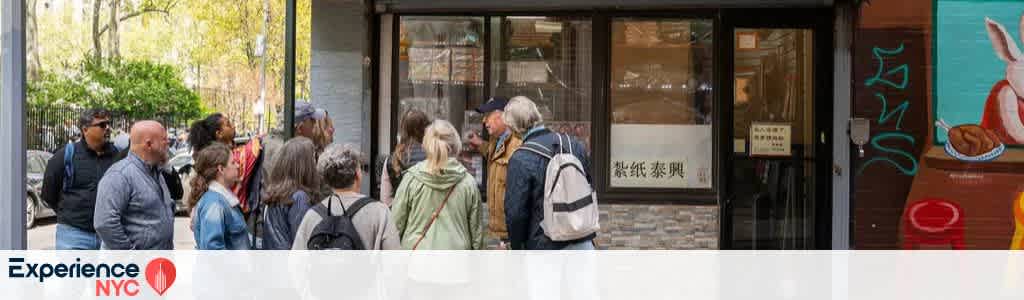 A group of people is gathered in front of a storefront with a cultural mural on the right, featuring vibrant colors and imagery. They appear to be on a guided tour in an urban setting, with trees visible in the background suggesting a city park nearby. The scene evokes a sense of exploration and community engagement within a diverse cityscape.