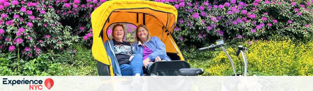Two people are enjoying a pedicab ride, seated under a bright yellow canopy. They are smiling, and in the background, vibrant purple and yellow flowers create a colorful scene. The image promotes the joy of exploring and experiencing NYC in a unique way.