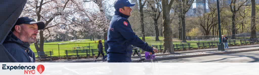 Two individuals are enjoying a sunny day in a park with trees and people in the background. One person is wearing a cap and looking ahead while the other is riding a bike. The park is lush with green grass and blooming cherry blossom trees. The atmosphere is lively with other visitors walking in the distance. A logo reading Experience NYC is visible in the foreground.