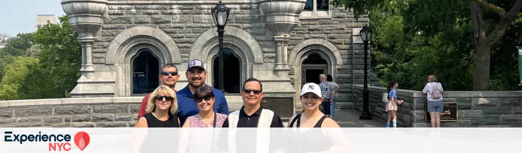 A group of six people stands on a stone terrace in front of a historic building with arched windows. They appear to be enjoying a sunny day. Tall trees and greenery are visible in the background. A walkway with visitors extends to the right. A logo at the bottom left reads Experience NYC.