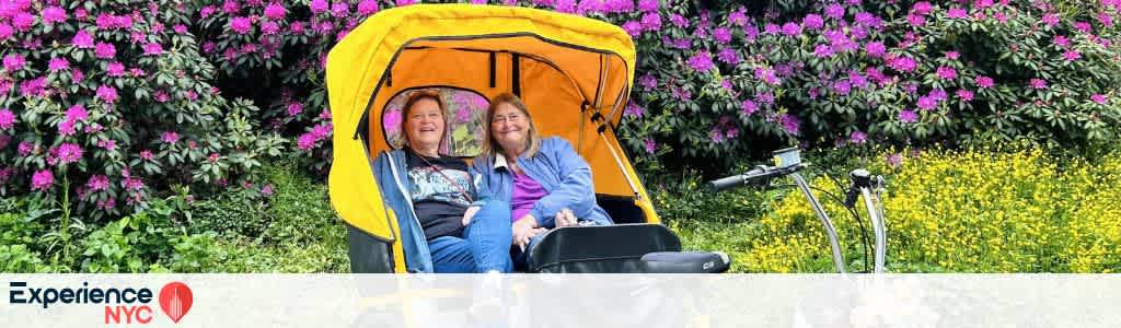 Two people are smiling while seated in a bright yellow pedicab. They're enjoying a ride through a park filled with vibrant purple and yellow flowers. A large lush bush forms a colorful backdrop. The words Experience NYC with a location pin icon appear in the bottom left corner.