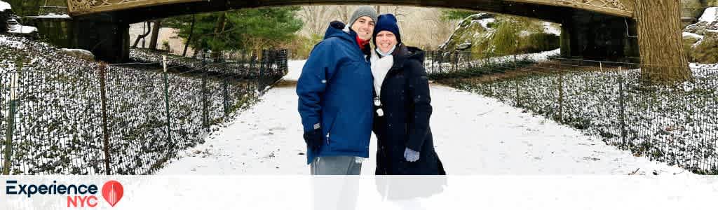 A couple stands smiling on a snow-covered path in a park, wearing winter clothing. Behind them, a decorative bridge and bare trees create a picturesque winter scene. The pathway is lined with a black metal fence partially covered with snow. The image promotes the beauty of a winter experience in NYC.
