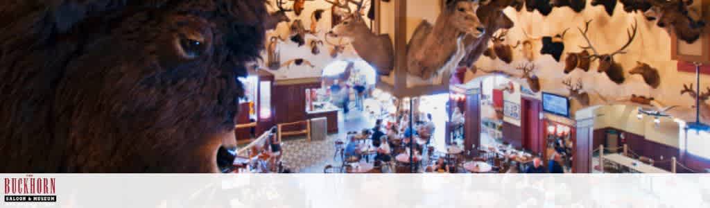 Interior view of a bustling restaurant with a rustic theme. The walls are adorned with numerous mounted animal heads, including deer and bison. Customers are seated at tables, enjoying their meals in a lively atmosphere. The room features warm lighting and wooden decor, creating a cozy and inviting environment. The Buckhorn Saloon and Museum logo is visible in the bottom left corner.
