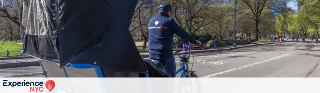 A person rides a pedicab along a wide, tree-lined road in a sunny urban park setting. The trees are lush with early spring foliage. A few pedestrians stroll in the distance. The pedicab has a black canopy, and the rider wears a dark jacket and cap. The scene suggests a leisurely, scenic ride in a vibrant city park environment. A logo with the words Experience NYC is shown at the bottom of the image.