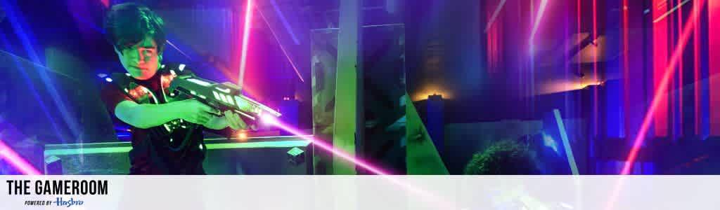 A person wearing a vest is engaged in a laser tag game, aiming a blaster while vibrant neon lights and colorful beams fill the room, creating an energizing and immersive atmosphere. The background showcases a mix of vivid pink, blue, and purple lights, enhancing the futuristic setting. Text reads: The Gameroom Powered by Hasbro.