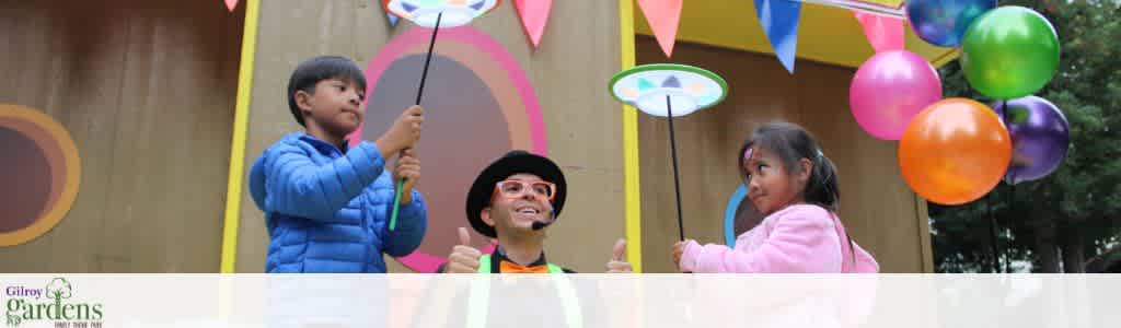Three children holding paper umbrellas and balloons at a colorful outdoor event