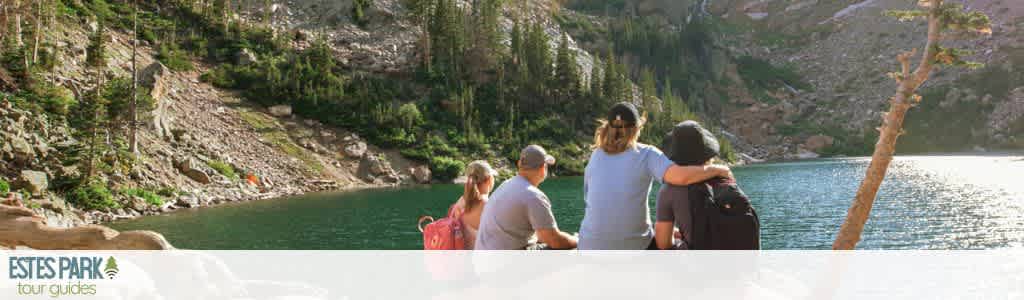 A group of people sits on rocks overlooking a serene mountain lake surrounded by evergreen trees and rocky slopes. The scene captures the tranquility of nature under a clear sky. This is an example of the scenic experiences offered by Estes Park tour guides, perfect for those seeking outdoor adventures and relaxation.