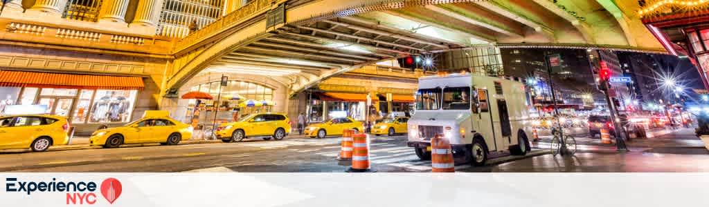 A nighttime city scene in New York City features a bustling street filled with yellow taxis, a white delivery truck, and bright traffic lights. The cityscape is vibrant with illuminated buildings and a lively urban atmosphere. An overpass stretches overhead, adorned with bright lights, capturing the essence of NYC's constant activity and energy. Orange traffic cones are visible on the street, contributing to the bustling city environment.