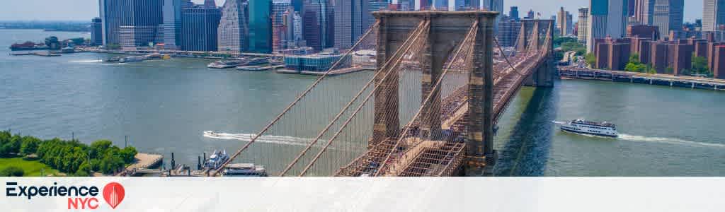 Aerial view of the Brooklyn Bridge leading into Manhattan, showcasing the city skyline with tall buildings against a clear blue sky. The East River flows beneath, with a boat cruising on the water. Lush greenery is visible on the left side. Text reads Experience NYC with a location pin icon.