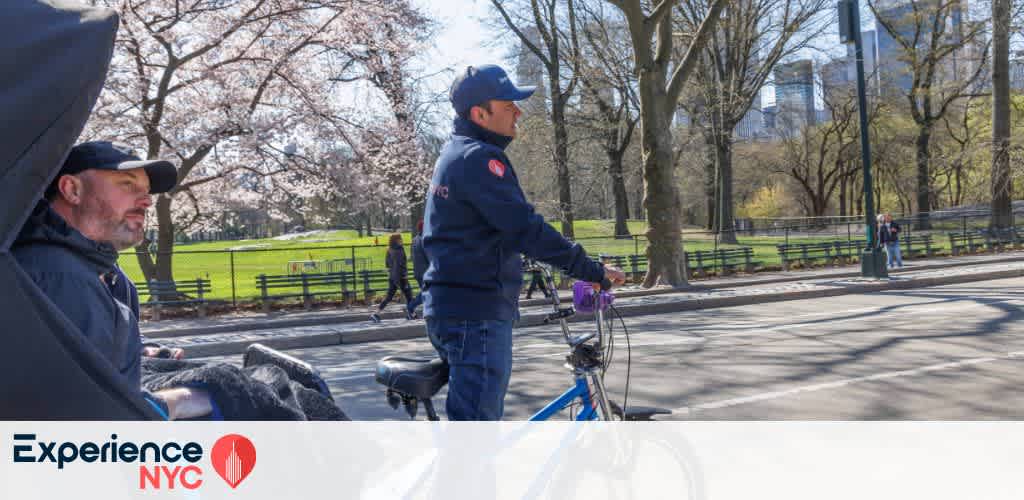 A pedicab rider in a blue uniform is cycling through a park with blooming trees and green lawns. A passenger sits comfortably in the back. The scene captures a sunny day in a bustling city park. The website logo Experience NYC is visible in the corner, suggesting an urban tour or travel experience.