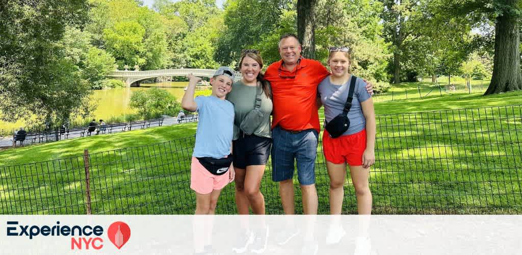 A family of four stands together smiling in a lush park setting. Behind them is a scenic view of trees and a lake with a stone bridge. The group is casually dressed, enjoying a sunny day. The scene conveys joy and relaxation. Experience NYC logo is at the bottom left corner of the image.
