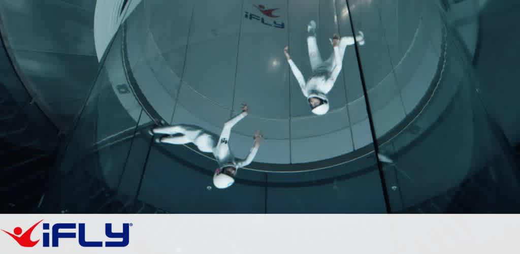 Two people in white flight suits and helmets are indoor skydiving in a vertical wind tunnel at iFLY. They are positioned horizontally, appearing to float, with their reflections visible on the glass walls. The environment is sleek and modern, showcasing an exciting and safe setting for skydiving enthusiasts of all skill levels. The iFLY logo is visible at the top and bottom of the image.