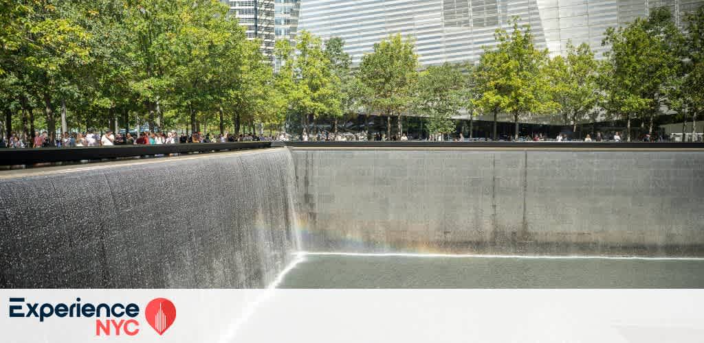 Image of the 9/11 Memorial in New York City featuring a large reflecting pool with cascading waterfalls along its dark stone walls. Surrounding the pool are lush green trees under a clear sky, with buildings in the background. Visitors are seen observing the solemn site. The Experience NYC logo is visible at the bottom left, showing an appreciation for cultural and historical exploration.