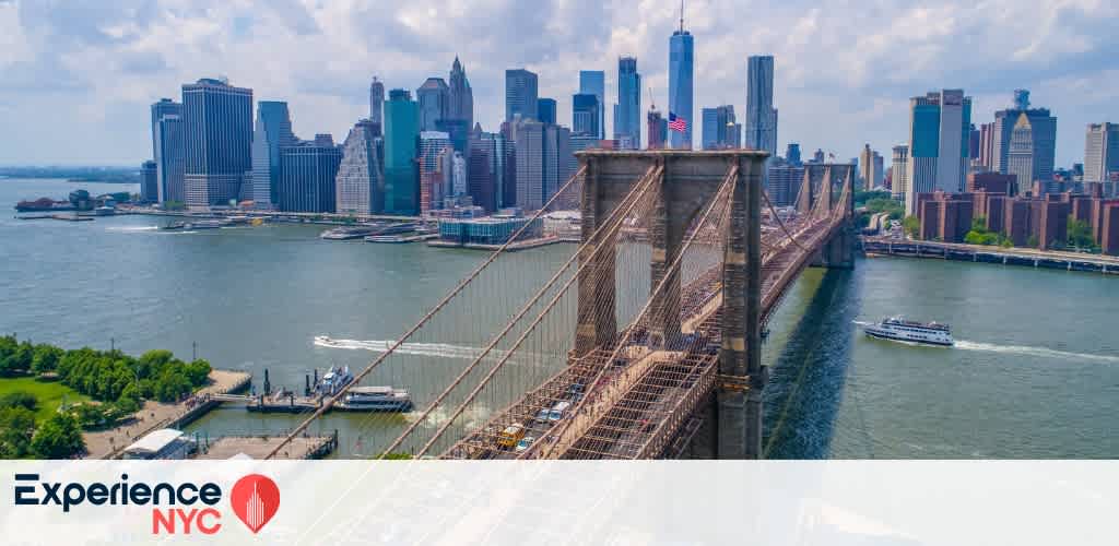 Aerial view of New York City showing the Brooklyn Bridge in the foreground and the Manhattan skyline in the background. The scene includes tall skyscrapers under a partly cloudy sky, with a river and a boat nearby. A green park and waterfront are visible on the left. The words Experience NYC are visible in the bottom left corner.