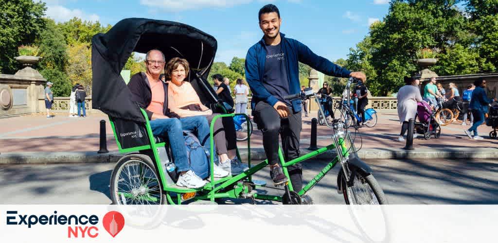 A smiling pedicab driver in a navy blue outfit enjoys a sunny day in a lively park setting. He is riding a bright green pedicab carrying two passengers, a man and a woman, both smiling. Visitors and trees fill the background, creating a cheerful atmosphere. Experience NYC branding is visible, enhancing the sense of leisure and adventure.