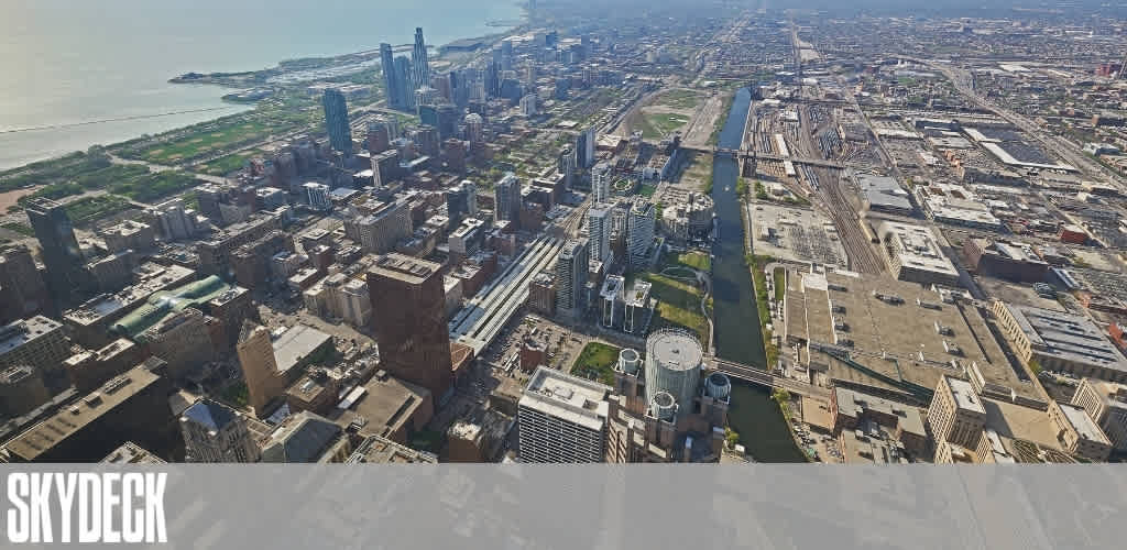 Aerial view of a cityscape featuring a mix of modern skyscrapers and older buildings. A river runs through the city, bordered by streets and green spaces. The sky is clear, and a large body of water is visible in the distance. The word Skydeck is prominently displayed in the lower-left corner.