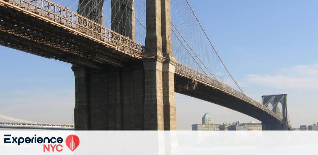 View of the Brooklyn Bridge stretching across the sky, with its iconic stone arches and cables. The blue sky provides a serene backdrop. Experience NYC logo is at the bottom with a stylized red landmark. City buildings can be seen in the distance, highlighting the urban setting of this famous New York City landmark.
