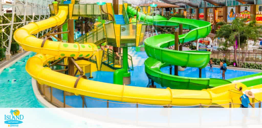 Colorful indoor water park featuring two twisting slides, one yellow and one green, ending in a pool. The setting includes a vibrant backdrop of shops and tropical plants. People are enjoying the facility, creating a lively and fun atmosphere suitable for families and friends seeking adventure and relaxation.