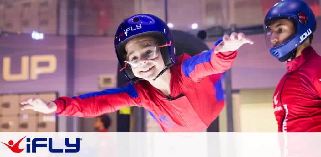 A child in red flight gear and a blue helmet is smiling while experiencing indoor skydiving at an iFLY facility. An instructor in similar gear guides them, ensuring safety. The background is softly blurred, highlighting the excitement of the activity. The iFLY logo is visible, emphasizing an adventurous experience for all ages.