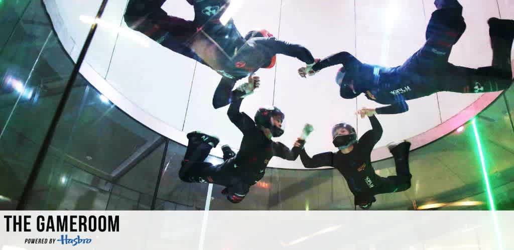 Four individuals in flight suits and helmets are skydiving indoors in a wind tunnel, forming a circle mid-air with arms linked. They are in a transparent chamber, creating an exciting and dynamic scene. The text The Gameroom powered by Hasbro is at the bottom, suggesting a thrilling activity or experience. The image conveys energy, teamwork, and innovation.