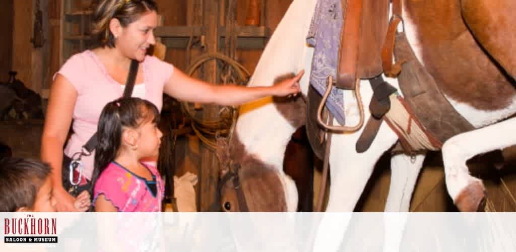 A woman and two children are exploring a museum exhibit. They are examining a brown and white horse mannequin with saddlery, showing interest and curiosity. The setting appears to be rustic, with wooden walls and artifacts. The Buckhorn Saloon and Museum logo is visible in the corner, indicating the location of the exhibit.