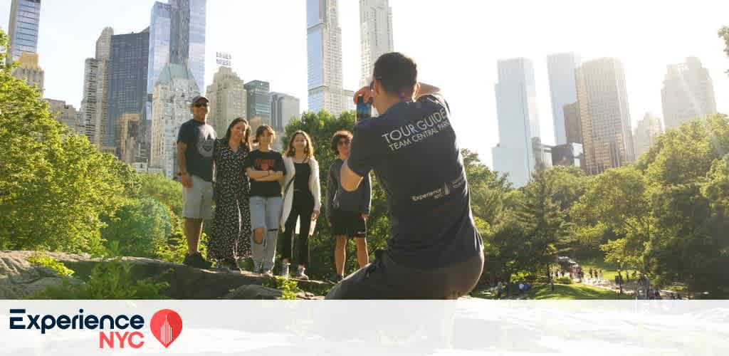 A group of people stands on a rock in a lush park setting with skyscrapers in the background. They are being photographed by a person wearing a tour guide shirt. The sun shines brightly, casting a warm glow on the scene. The foreground features green trees contrasting with the urban skyline. The image promotes an urban nature experience in NYC. The logo in the corner reads Experience NYC.